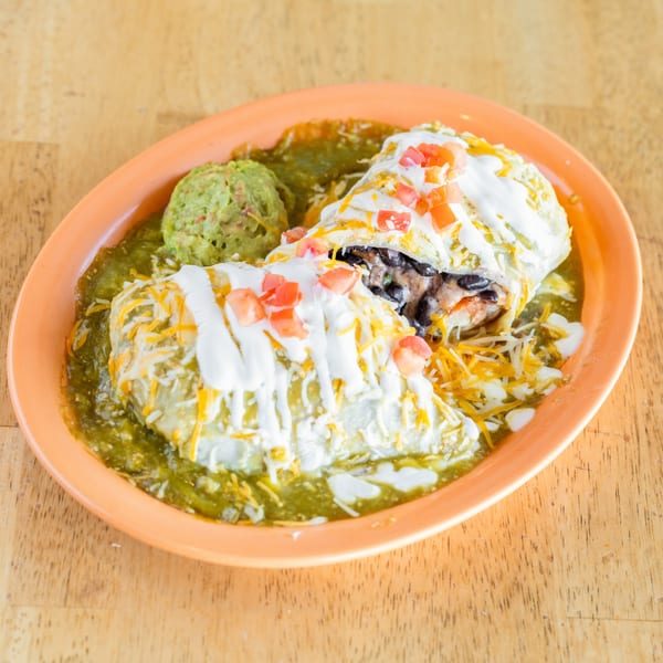 Two burritos covered in green chili sauce, cheese, sour cream, and diced tomatoes, with guacamole.